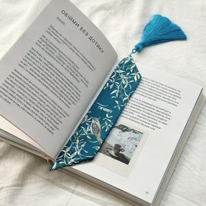 May include: A teal bookmark with a white floral pattern and a bird illustration rests on an open book. The bookmark has a blue tassel and the word "OBIMY" is printed on it.