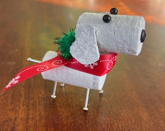 May include: A handmade dog figurine crafted from a repurposed cork, featuring black bead eyes and a black nose. It wears a red ribbon with white snowflake accents and a green pom-pom collar. The dog stands on wire legs.