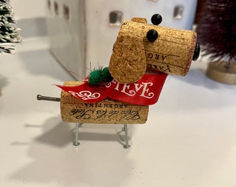 May include: A handcrafted dog figurine made from a wine cork, featuring black bead eyes, a red ribbon collar with the name "Steve", and screw legs. The dog is part of a holiday decoration set.