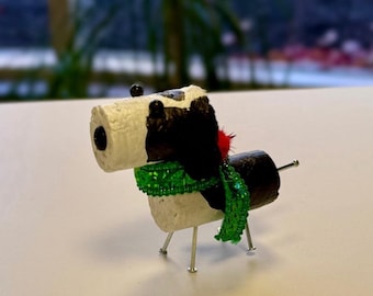 May include: A small dog figurine crafted from two corks, featuring a white head with black painted ears and a black body. The dog wears a green sequined scarf, has black bead eyes, a red pom-pom on its back, and stands on four silver metal pins.