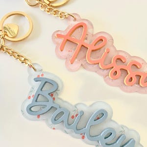 May include: Two personalized acrylic keychain charms with names "Alissa" and "Bailey" in cursive script. The charms are layered with a floral pattern and have a gold chain and clasp.