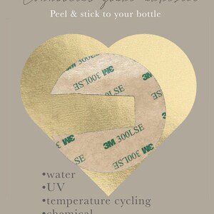 May include: Close-up of two round, brown adhesive pads with the text "3M 300LSE" printed on them. The adhesive pads are on a gold heart-shaped background. The text "Commercial grade adhesive" and "Peel & stick to your bottle" is at the top of the image. The text "water, UV, temperature cycling, chemical resistance" is below the adhesive pads. The text "*it will be visible on clear and transparent acrylic sheets" is at the bottom of the image.