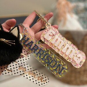 May include: Two personalized keychains with names "Emma" and "Harrison" written on them. The Emma keychain is pink and white with glitter, while the Harrison keychain is yellow and blue with a pattern of small dinosaurs.