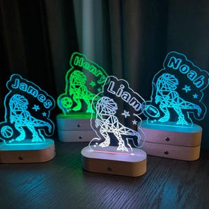 May include: Four personalized night lights featuring a geometric dinosaur design with a basketball. Each light has a different name on it: Henry, James, Liam, and Noah.