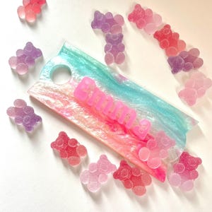 May include: A rainbow-colored resin tray with the name "Emma" spelled out in pink letters. The tray is surrounded by small, glittery gummy bear charms in various shades of pink, purple, and red.