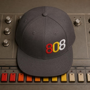 May include: A charcoal gray baseball cap with a flat brim and a structured crown. The front of the cap features the embroidered number 808, with the 8 in red, the 0 in yellow, and the 8 in white. The cap has a button on top and stitched eyelets.