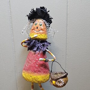 Könnte beinhalten: Eine skurrile Halloween-Dekoration in Form einer Candy-Corn-Hexe. Die Figur hat ein lächelndes Gesicht, einen schwarzen Hut und hält einen kleinen eulenartigen Korb. Der Sockel ist grün mit silbernem Lametta.