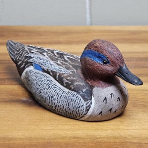Op de afbeelding: Een handgesneden houten eendenbeeldje. De eend heeft een bruine kop met een blauwe streep, een zwarte snavel en een lichaam met grijze, bruine en witte details. De eend rust op een houten oppervlak.