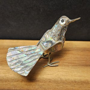 May include: A decorative bird figurine with iridescent shell-like detailing on the tail and body. The bird has a long beak and is perched on wire legs. The figurine is set against a dark background and rests on a wooden surface.