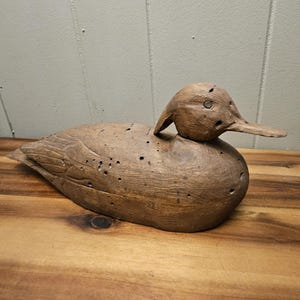 May include: A wooden duck decoy, carved from light brown wood. The duck has a detailed head, beak, and body, with small holes throughout. The decoy is placed on a wooden surface.