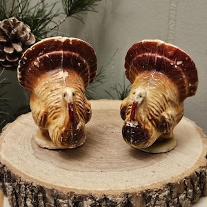 May include: Two ceramic turkey figurines with brown, tan, and red coloring. The turkeys have detailed feathering and stand on small bases. They are displayed on a wooden slice, with a pine cone and greenery in the background.