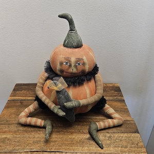 May include: A fabric pumpkin doll with a painted face, featuring a green stem, orange body with vertical lines, and a black ruffled collar. The doll has long, striped arms and legs, and is holding a small dark object tied with a yellow ribbon.