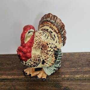 Puede incluir: Una figura de pavo de cerámica con una carúncula y un pico de color rojo brillante, con el cuerpo pintado en tonos crema, marrón y negro. El pavo tiene una cola en abanico con marcas de plumas detalladas y patas texturizadas con garras negras.