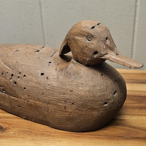 May include: A carved wooden duck decoy, featuring a detailed head and body. The duck is a natural brown color with visible wood grain and small holes. The decoy is resting on a wooden surface.