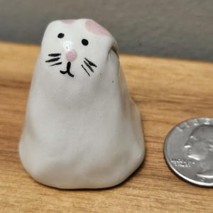 Op de afbeelding: Een kleine, witte keramische kattenfiguur met roze accenten op het hoofd. De kat heeft zwarte ogen en snorharen, en een roze neus. Een munt ligt naast het beeldje ter vergelijking. Op een houten oppervlak.