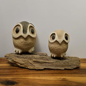May include: Two ceramic owl figurines, one large and one small, perched on a piece of driftwood. The owls have tan bodies, dark eyes, and detailed facial features. The larger owl has a dark gray head. The driftwood is a light brown color.