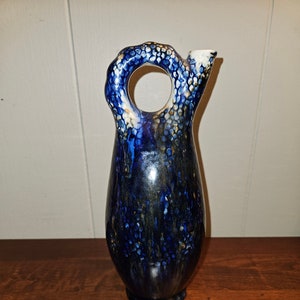 May include: A blue and gold ceramic pitcher with a unique, textured, and abstract design. The pitcher has a wide, round opening and a handle that forms a circle.