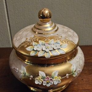 May include: A white ceramic jar with a gold lid and floral designs. The jar has a round body and a wide mouth. The lid has a gold knob and a gold band around the top. The floral designs are painted in gold and white.