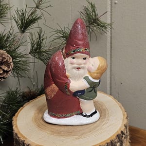 May include: A ceramic Santa Claus figurine in a burgundy robe and hat, embracing a child in a green and white outfit. The figurine has a white beard and is on a wooden slice base, with pine branches in the background.