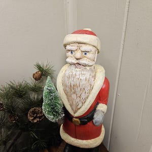 May include: A wooden Santa Claus figurine with a red coat, white trim, and a long white beard. The figurine is holding a small green Christmas tree and stands on a dark wooden base. The Santa has blue eyes and a red hat.