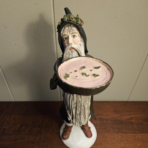 May include: A figurine of a bearded man wearing a black robe and a holly wreath, holding a bowl of pink pudding with holly sprigs on top.