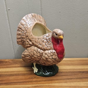 May include: A ceramic turkey planter with a tan body, red wattle, and yellow beak. The turkey has a large tail and sits on a green base. The planter has an opening on its back for plants or decorations.