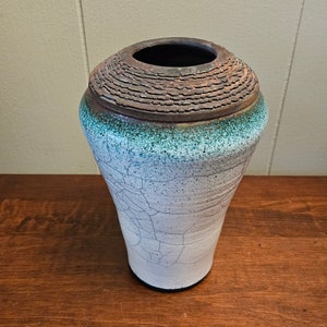 Może przedstawiać: Biały ceramiczny wazon z pękniętą glazurą i brązową krawędzią. Wazon ma turkusowy pasek wokół górnej części.