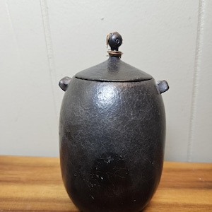 May include: A dark gray, hammered metal jar with a lid and small handles. The jar has a rounded shape and a textured surface. The lid is topped with a small, decorative knob. The jar sits on a wooden surface.