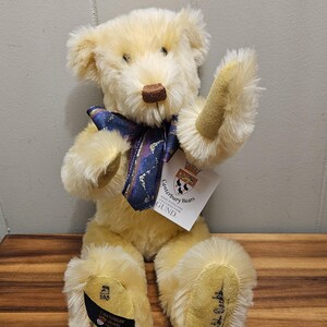 May include: A plush, cream-colored teddy bear with a dark brown nose and a blue and purple patterned bow tie. The bear has a tag that reads "Canterbury Bears" and "GUND". The bear is sitting on a wooden surface.