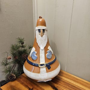 May include: A handcrafted Santa Claus figurine with a brown hat and coat, white beard, and blue gloves. The ceramic figure has a blue belt with a gold buckle and is displayed on a wooden surface. The Santa Claus figurine is a festive holiday decoration.