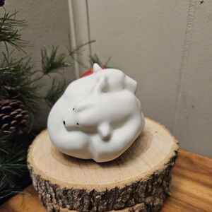 Könnte beinhalten: Eine weiße Keramik-Eisbärfigur mit roter Mütze, die auf einer Holzscheibe ruht. Der Bär hat schwarze Augen und ist zusammengerollt. Die Figur steht auf einer rustikalen Holzscheibe, mit einem Holzuntergrund im Hintergrund. Das Gesamtthema ist Winter und Weihnachten.