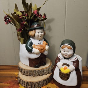 May include: Two ceramic Thanksgiving planter figurines. One figure is a man holding a turkey, the other is a woman holding a basket of fruit. The planters are white with brown and green accents. The man planter is on a wood slice base.