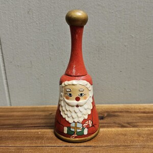 May include: A wooden Santa Claus bell with a red and white painted design. The bell has a gold top and a white beard. Santa is holding a gift and a candy cane.