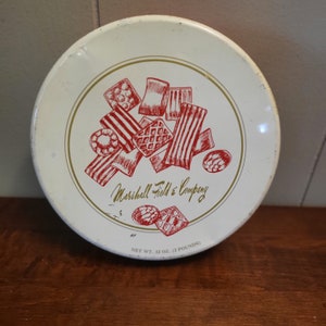 Op de afbeelding: Een witte ronde blik met een rood en gouden ontwerp. De blik heeft een tekening van verschillende koekjes en de tekst "Marshall Field & Company". De onderkant van de blik heeft de tekst "Net Wt. 32 oz. (2 pounds)".
