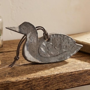 May include: A flat, gray, metallic duck-shaped ornament with a textured surface and a dark leather cord looped through a hole near the neck. The duck figure features a pointed beak, a rounded head, and a stylized wing detail on its side.