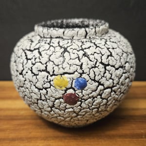 May include: A ceramic vase with a white, cracked glaze and a black rim. The vase features a face design with yellow, blue, and red dots. The vase is sitting on a wooden surface.