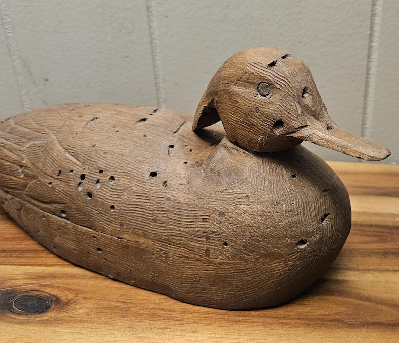 May include: A carved wooden duck decoy, featuring a detailed head and body. The duck is a natural brown color with visible wood grain and small holes. The decoy is resting on a wooden surface.