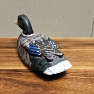 Puede incluir: Figura de pato de madera tallada a mano. El pato tiene una cabeza negra, un cuerpo gris y blanco con detalles azules en las alas y un pecho blanco. El pato est&aacute; colocado sobre una superficie de madera.