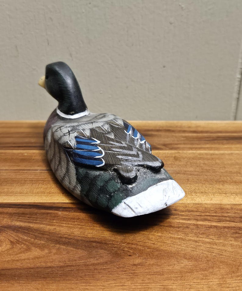 Puede incluir: Figura de pato de madera tallada a mano. El pato tiene una cabeza negra, un cuerpo gris y blanco con detalles azules en las alas y un pecho blanco. El pato est&aacute; colocado sobre una superficie de madera.