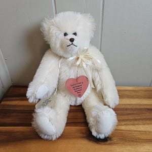 May include: A white teddy bear with a cream-colored bow around its neck. The bear has a heart-shaped tag that reads "Whitney An Original Cranberry Mountain Bear". The bear is sitting on a wooden surface.