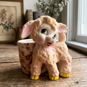 May include: A ceramic planter shaped like a lamb with textured light pink wool, large expressive eyes, and bright yellow hooves. The lamb stands beside a brown, woven-style basket container, featuring pink inner ears and a small red mouth.