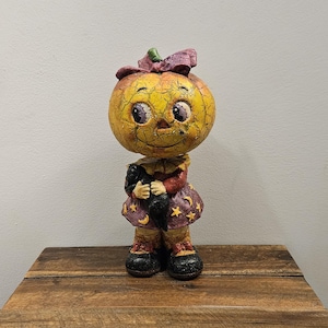 May include: A decorative figurine featuring a pumpkin-headed character with a cracked yellow surface, wearing a purple dress adorned with yellow stars and moons, a purple bow, and black shoes, while holding a small black cat in its arms.