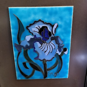Puede incluir: Un azulejo de cerámica azul y blanco con una imagen pintada a mano de una flor de iris con hojas negras y verdes. El azulejo está firmado "SK80" en la esquina inferior derecha.