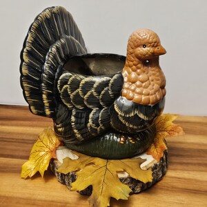 May include: A ceramic turkey-shaped planter with a brown and black body, a detailed tail, and a brown head. The planter sits on a wooden base with yellow and orange fall leaves, perfect for seasonal decor.