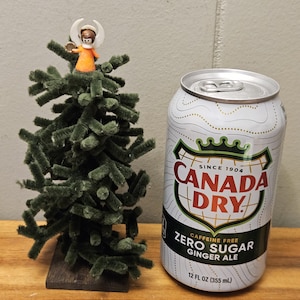 May include: A small, handmade Christmas tree made of green pipe cleaners, with an angel figurine on top. Next to it is a 355 ml can of Canada Dry Zero Sugar Ginger Ale.