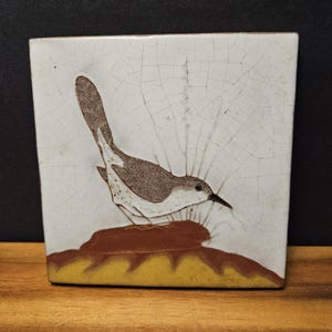 Puede incluir: Un azulejo de cerámica cuadrado con un pájaro pintado a mano en marrón y blanco, posado sobre un montículo marrón encima de una base amarilla. El fondo blanco tiene un acabado de esmalte craquelado con una red de líneas finas.