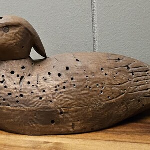 May include: A weathered, wooden duck decoy with a detailed carved head and body. The duck is painted in shades of brown and has numerous small holes throughout its body, giving it a rustic appearance. The decoy is resting on a wooden surface.