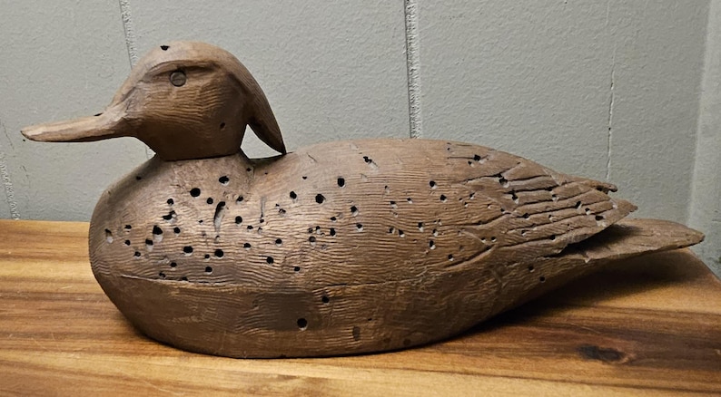 May include: A weathered, wooden duck decoy with a detailed carved head and body. The duck is painted in shades of brown and has numerous small holes throughout its body, giving it a rustic appearance. The decoy is resting on a wooden surface.