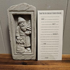 May include: A gray concrete sculpture of Santa Claus holding a Christmas tree and a bag of gifts. The sculpture is framed in a rectangular opening in a gray concrete wall. The sculpture is a Carruth Original, as indicated by the text on the card next to the sculpture. The card reads "Thank You for Choosing a Carruth Original. Every piece we create portrays the satire and whimsy that is so unique to every Carruth Original's work. We hand pour every Carruth original using the highest quality, most durable materials available. Which means one of the many personality traits of your piece may include the occasional minor surface blemish. These tiny flaws add individuality to your creation and have no effect whatsoever on its durability. Of course, our materials were also chosen because they quickly acquire their own natural patina, making your casting even more unique. But if the normal effects of nature stain your artwork beyond your own individual tastes, just do what we do - scrub it with a brush and a little detergent or bleach. We sincerely hope your original brings you years of pleasure. It was designed to do just that."