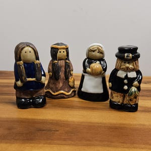 Puede incluir: Cuatro figuras de cerámica que representan figuras históricas. Las figuras incluyen un nativo americano, una mujer con un vestido largo y un peregrino. Las figuras están pintadas en varios colores, incluyendo azul, marrón, negro y blanco. Las figuras están sobre una superficie de madera.